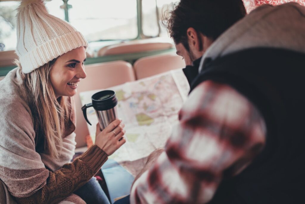 A smiling woman in a knit beanie holds a travel mug while talking to a man in a camper van.