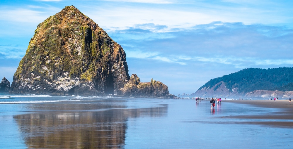 A large sea stack, Haystack Rock, rises from a sandy beach on the Oregon coast with calm, reflective water.