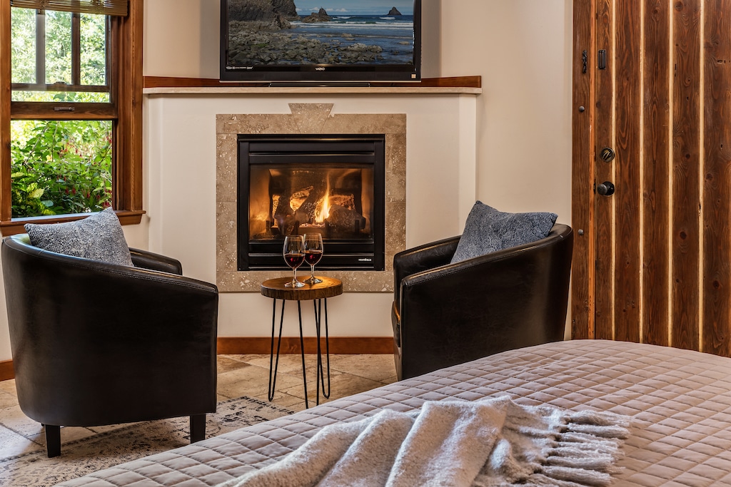 Two armchairs face a corner fireplace with a TV above it, featuring a small table with two wine glasses between them.