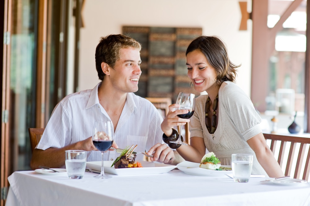 A couple sits at an outdoor table with white tablecloths, eating dinner and drinking red wine.