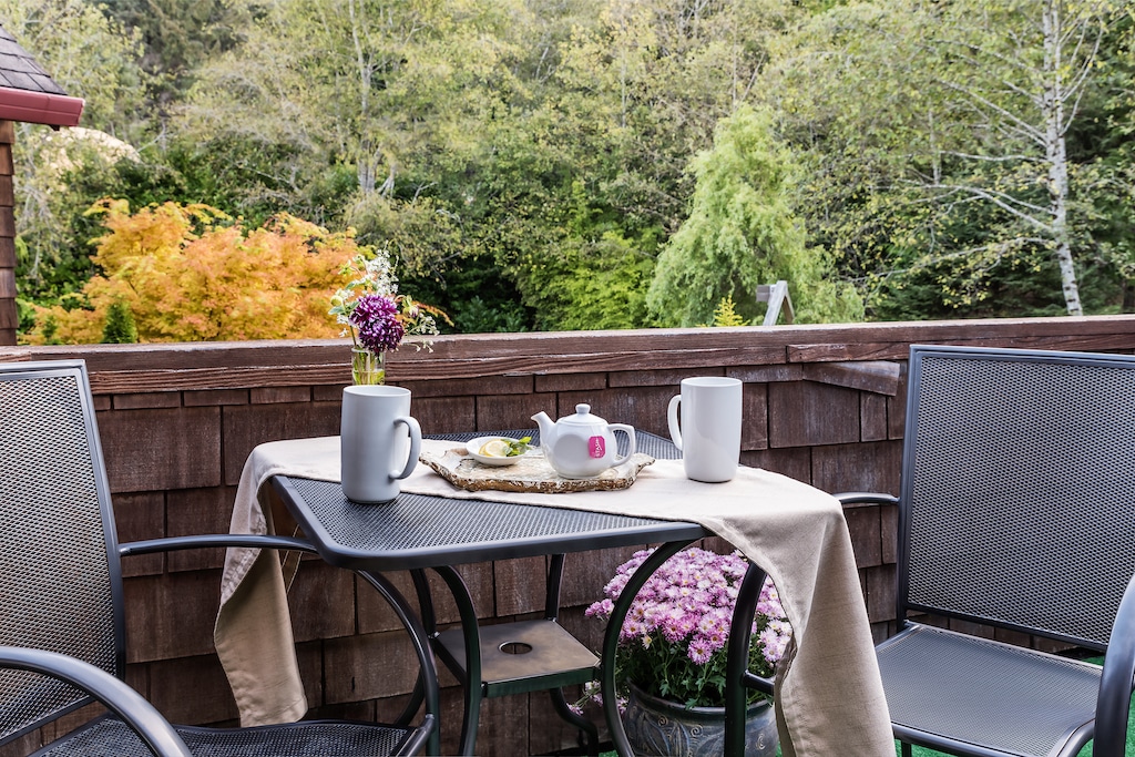 A small table with a teapot and two mugs on a balcony, overlooking a lush garden with trees.