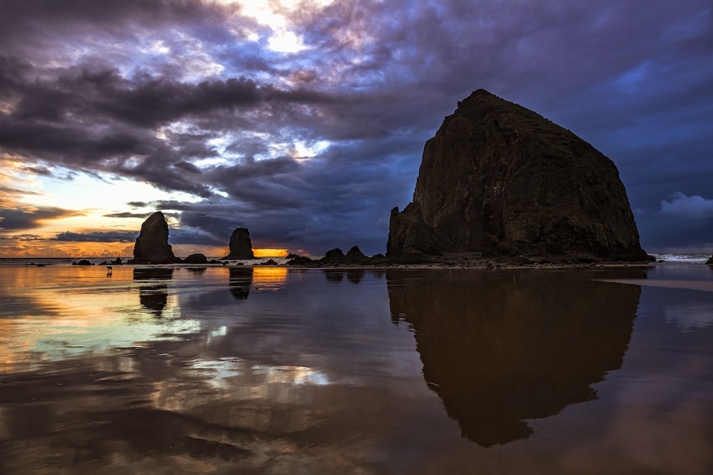 Haystack Rock at sunset, with its massive silhouette reflected in the wet sand on the beach.