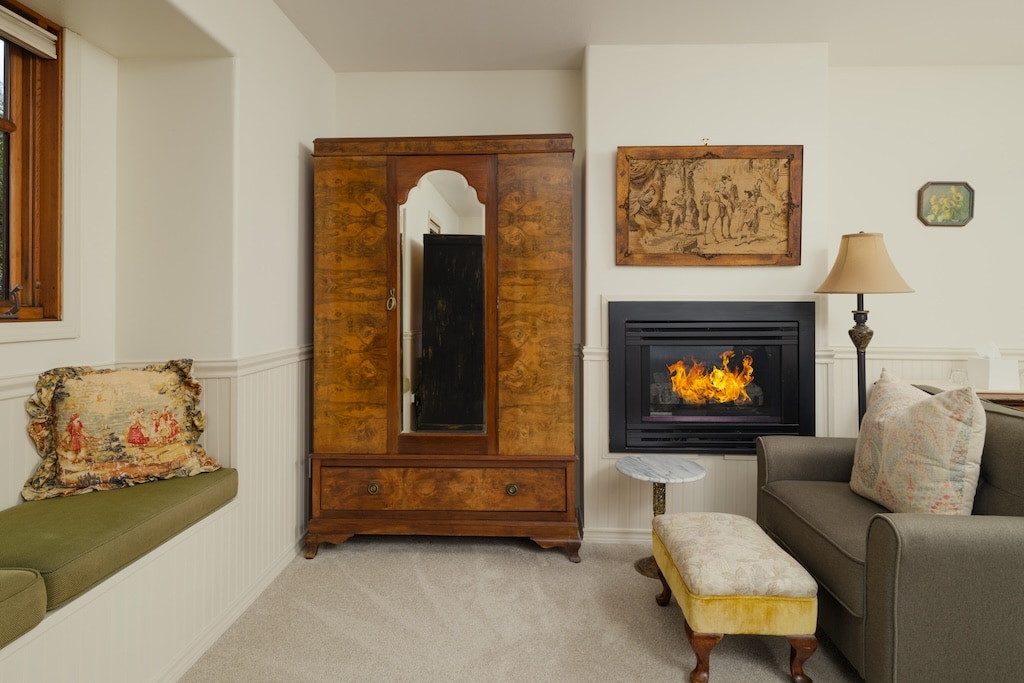 A room with a wooden wardrobe, a fireplace with a lit fire, a sofa, a stool, and a window seat with a patterned pillow.