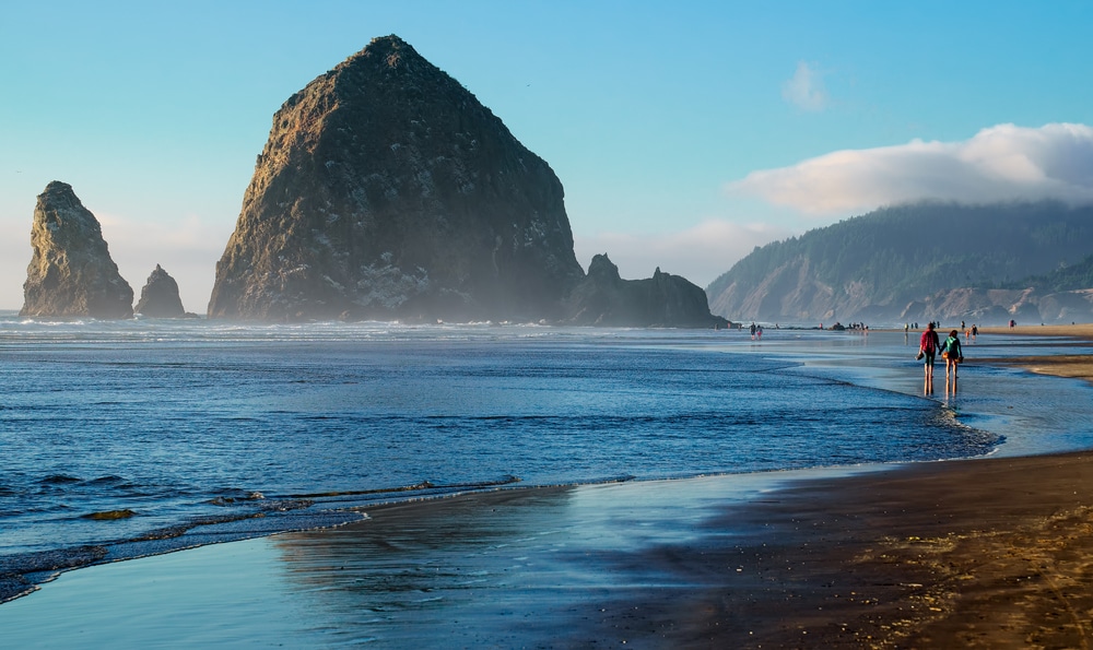 A wide shot of Haystack Rock on an Oregon beach, with ocean waves washing onto the sandy shore at sunset.