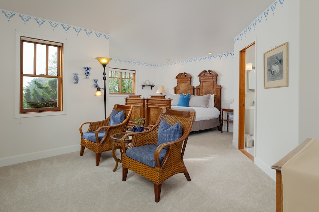 A bedroom with two wicker armchairs, a small table, a queen bed, and light-colored walls with blue decorative trim.