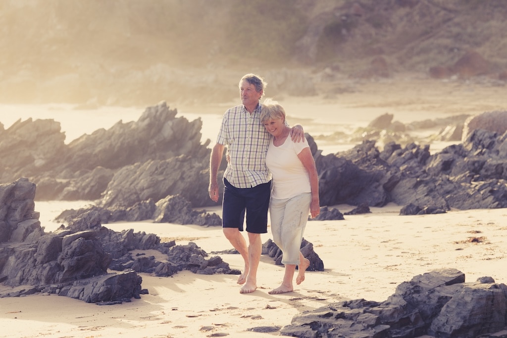 An elderly couple walks arm in arm across a rocky beach during a sunlit afternoon.