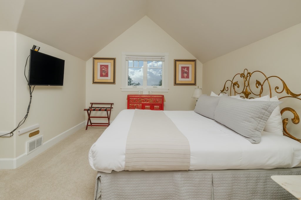 A hotel-style bedroom with a queen bed, mounted television, window, and luggage rack under a sloped ceiling.