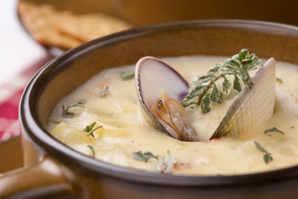 A bowl of creamy clam chowder garnished with a whole clam and herbs, with crackers in the background.