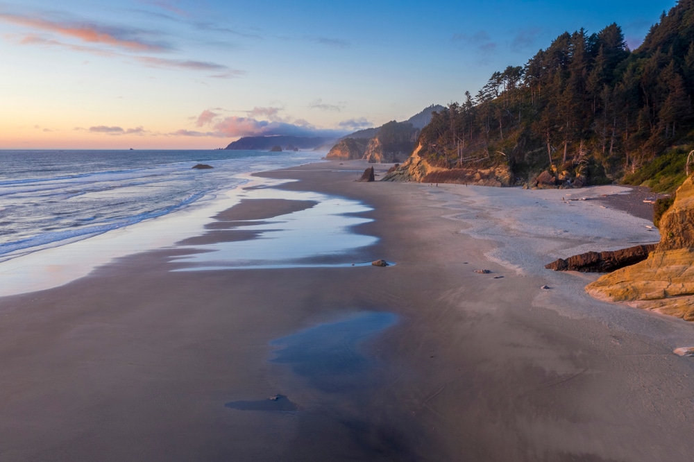 A sandy beach at twilight with calm waves, tide pools, and forested cliffs along the shoreline.