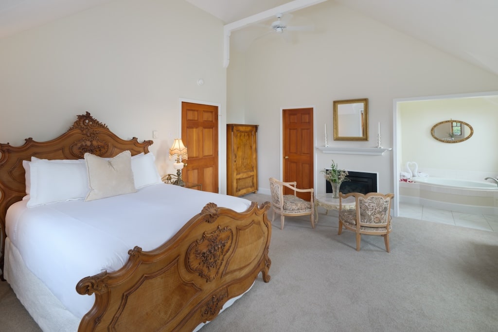 A spacious, high-ceilinged bedroom with a carved wood bed, fireplace, seating area, and a view into an adjacent bath.