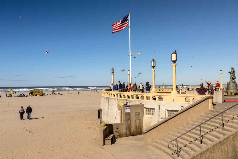 People walk on the beach near a stone observation deck flying an American flag under a clear blue sky.
