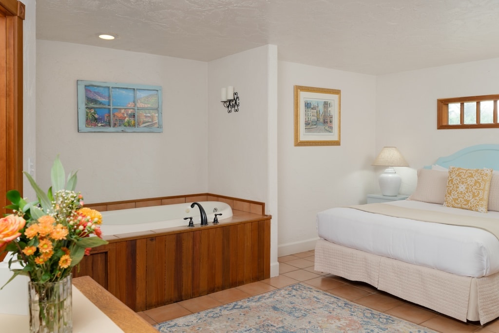 A bedroom with a wooden bathtub, a double bed, white walls, and a bouquet of flowers in the foreground.