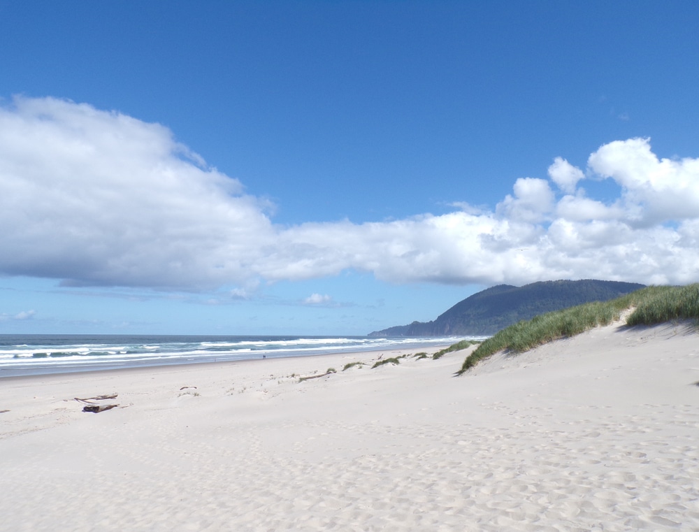 A sandy beach with dunes on the right, ocean waves, and mountains in the distance under a blue sky with clouds.