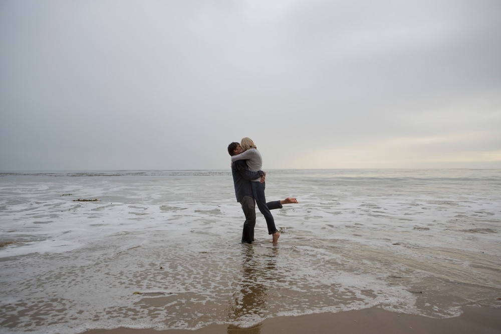 A couple kisses and embraces in the shallow water at the beach under an overcast sky.