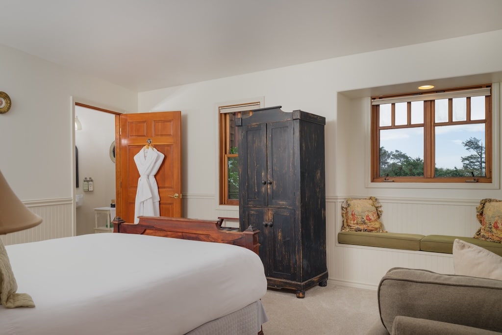 A bedroom with a white bed, a dark cabinet, an open door with a robe, and a window seat.