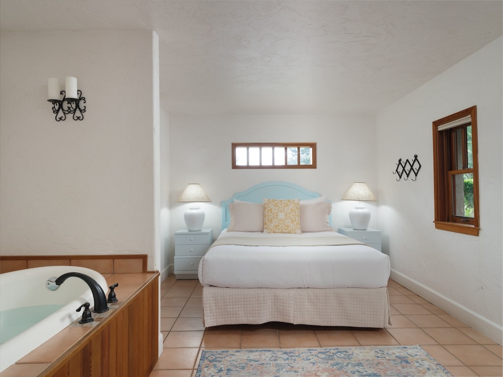 A hotel room featuring a bed with a blue headboard, flanked by bedside lamps, and an adjacent soaking tub in the front.