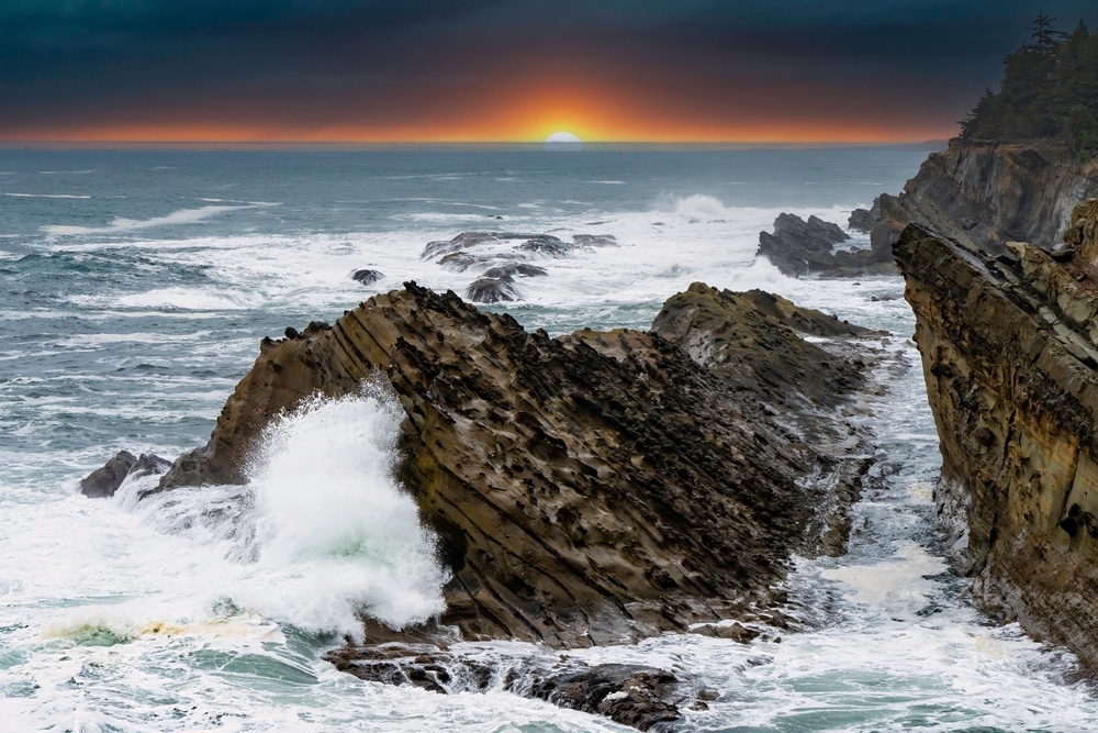 Waves crash against jagged, layered rock formations along a rugged coastline under a dark, orange-lit sunset horizon.