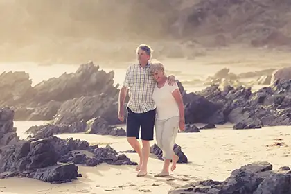 An older couple walks arm-in-arm along a sandy beach near dark rocks at sunset.