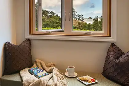 A window seat with two brown pillows, a book, a blanket, a cup of coffee, and a small plate of food.