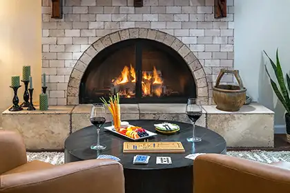 A cozy living room featuring a brick fireplace with a fire, two chairs, and a round table with wine and snacks.