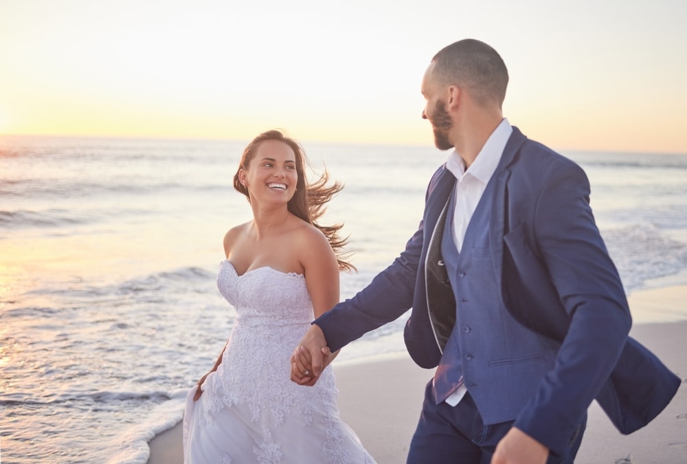 A bride in a white gown and a groom in a blue suit hold hands while walking along a beach at sunset.
