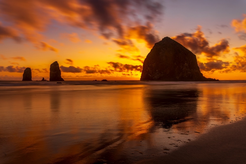 A dramatic orange sunset over Haystack Rock in Cannon Beach, Oregon, with reflections on the wet sandy beach.