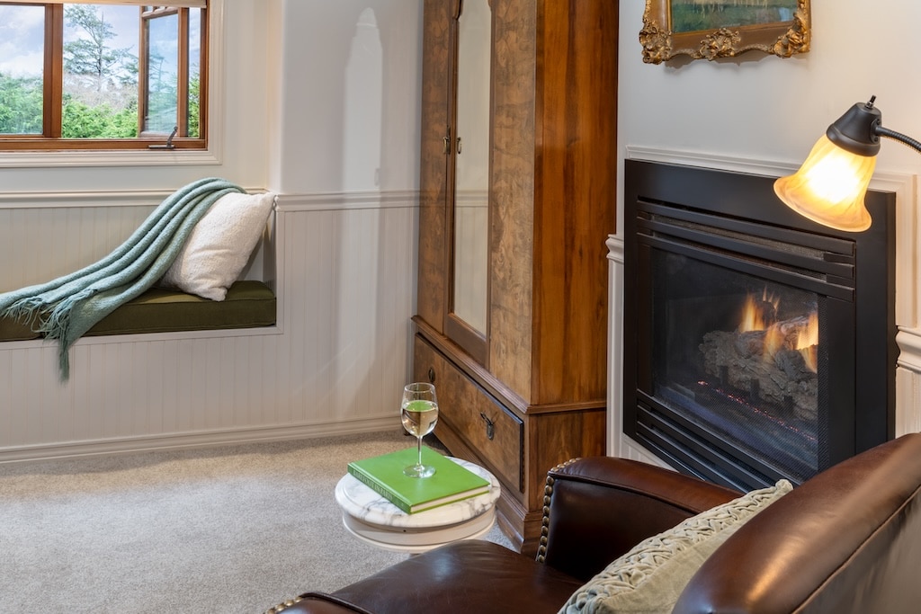 A cozy room with a window seat, a fireplace, a wooden wardrobe, and a leather armchair with a side table.