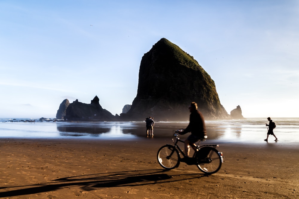 A person rides a bicycle along a sandy beach near Cannon Beach, Oregon, with Haystack Rock in the background.