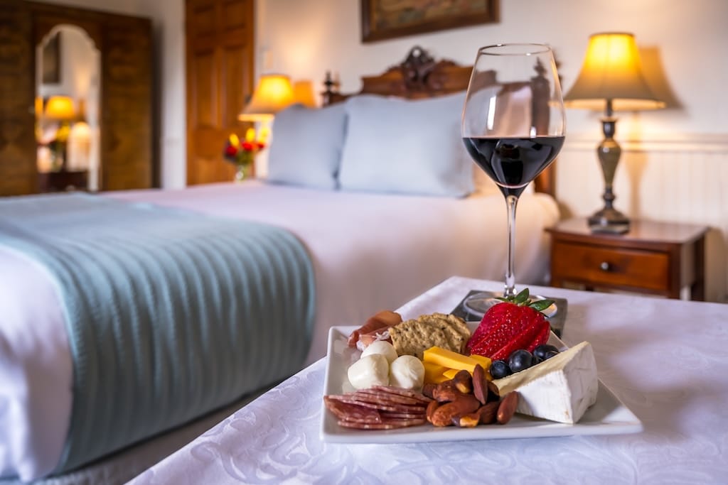A plate of snacks and a glass of red wine on a bed in a cozy bedroom with lamps and a made bed in the background.