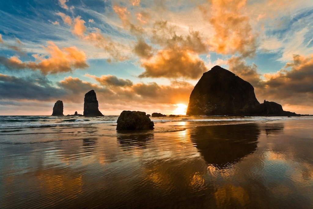 Sea stacks on a sandy beach at sunset, with golden clouds reflecting in the wet sand at low tide.