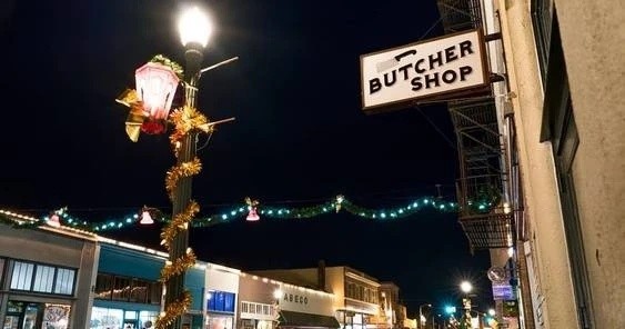 A street at night featuring a "Butcher Shop" sign, a decorated lamp post, and festive lights against a dark sky.