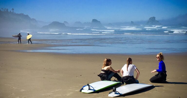 Three people sit on a sandy beach with surfboards, while two others walk along the shore in the background.