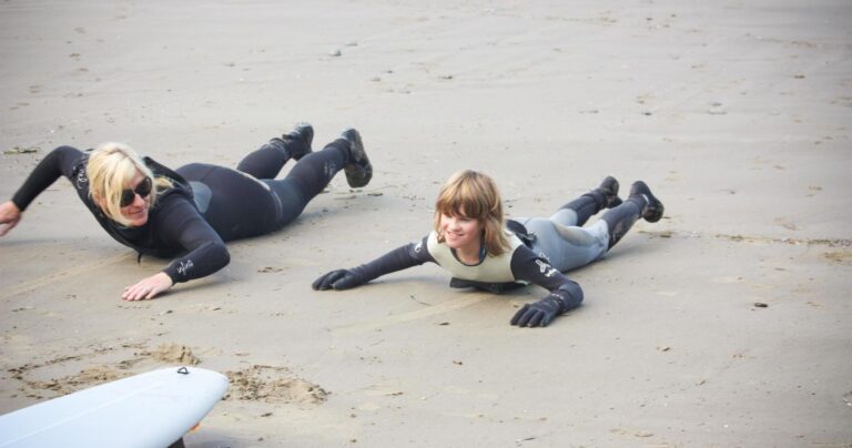Two people in wetsuits lie prone on a sandy beach, practicing surfing maneuvers.