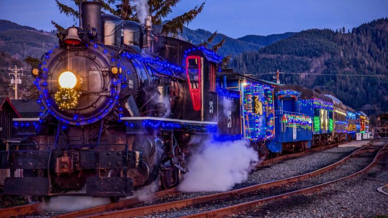 A steam train decorated with blue Christmas lights travels along tracks at dusk, emitting steam.