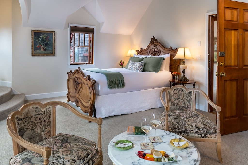 A hotel room with a queen bed, two patterned armchairs, a marble coffee table, and a wooden door.