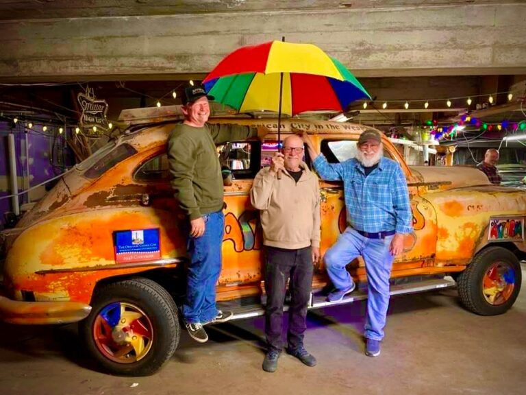 Three men stand beside an orange, vintage, decorated car, one holding a large multi-colored umbrella.