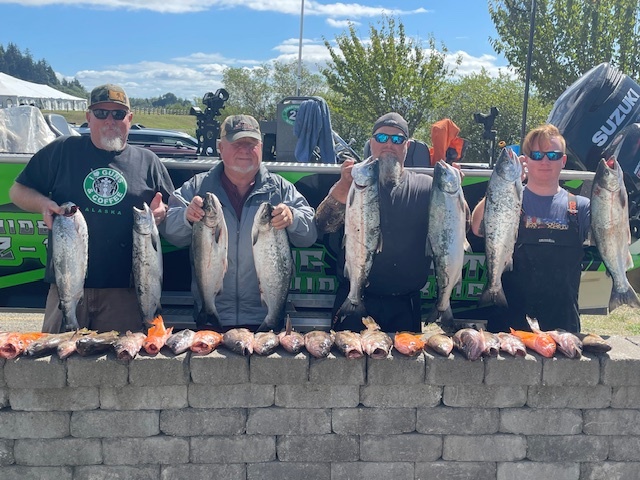 Four people stand behind a brick wall, each holding a large salmon in front of a boat, with more fish on the wall.
