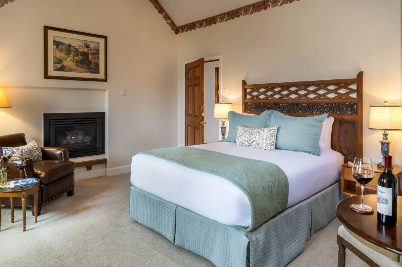A hotel room with a queen bed, a fireplace, leather chair, a side table with wine, and a framed picture on the wall.