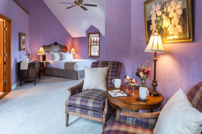 A bedroom with lavender walls, a bed in the back, and two plaid armchairs around a small table in the foreground.