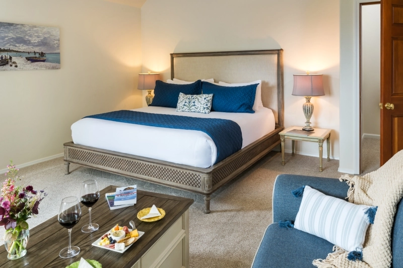 A hotel room features a king bed with blue accents, a wooden coffee table with wine, and a blue armchair.