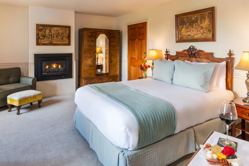 A hotel room with a queen bed, fireplace, armchair, ottoman, wardrobe, and a snack platter with wine in foreground.