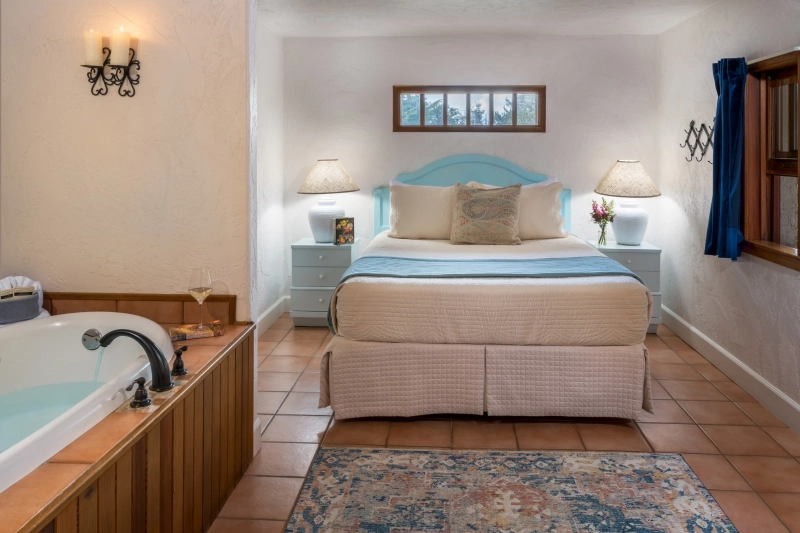 A bedroom with a queen bed, two nightstands, a soaking tub in the foreground, and tile flooring.
