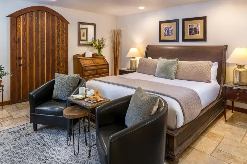 A furnished bedroom with a large brown bed, two leather armchairs, a wooden desk, and a rustic arched wooden door.