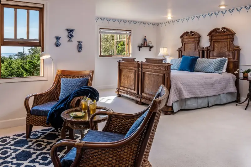 A bedroom with two wicker chairs, a small table, a large wooden bed, and windows overlooking greenery.