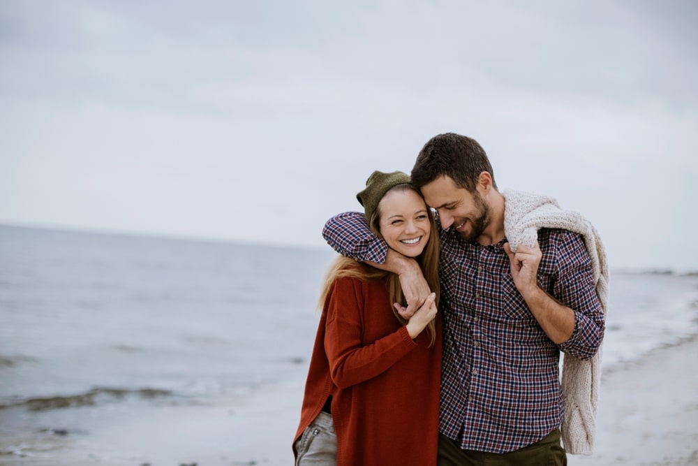 A happy couple stands on a beach, with the man embracing the woman from behind.