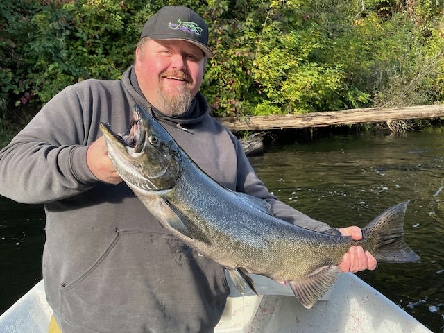 A man in a baseball cap and grey hoodie holds a large salmon while sitting in a small boat on a river.